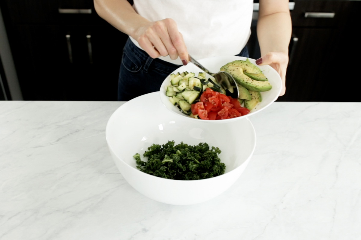 Adding tomatoes and cucumbers to a bowl of massaged kale to make the wrap filling and leaving the avocado on the side.